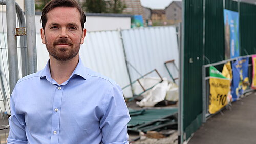 Adam Harley standing in front of an area partly fenced off with rubble and other construction materials in a disorganised fashion.