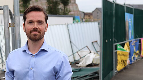 Adam Harley standing in front of an area partly fenced off with rubble and other construction materials in a disorganised fashion.