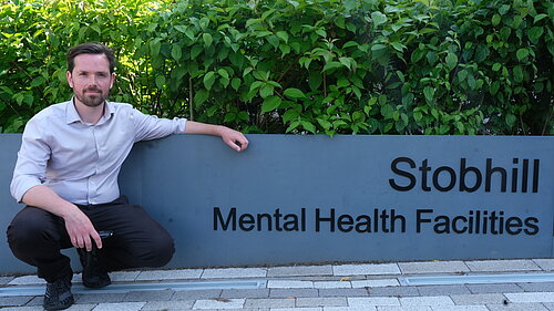 Adam Harley bending down next to the Stobhill Mental Health Facilities sign. 