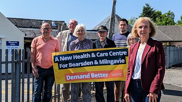 Susan Murray in front of a group of campaigners who are holding up a banner that reads "A new health centre for Bearsden and Milngavie. Demand Better. Scottish Liberal Democrats".