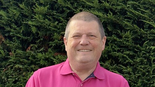 Photo of Ben Langmead smiling in a pink polo top with a hedge behind him.