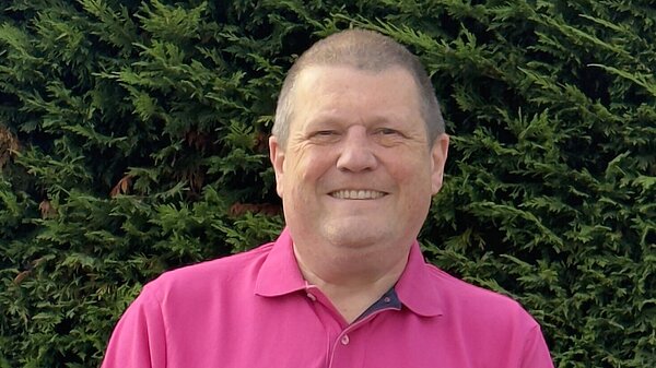 Photo of Ben Langmead smiling in a pink polo top with a hedge behind him.