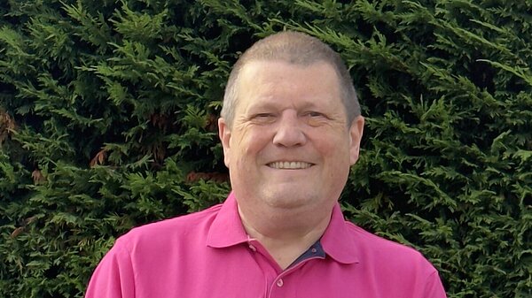 Photo of Ben Langmead smiling in a pink polo top with a hedge behind him.