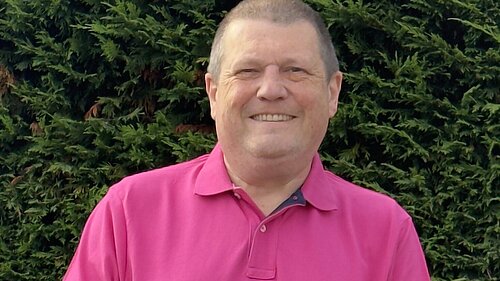 Photo of Ben Langmead smiling in a pink polo top with a hedge behind him.