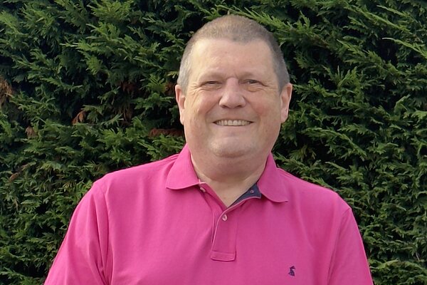 Photo of Ben Langmead smiling in a pink polo top with a hedge behind him.