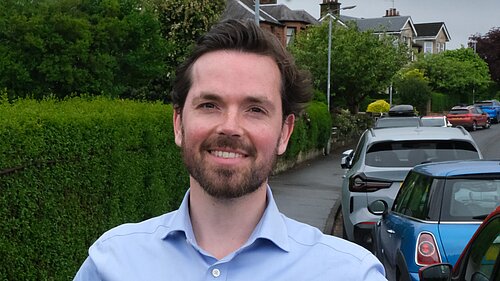 Adam Harley standing smiling with cars parked on the road next to the pavement behind