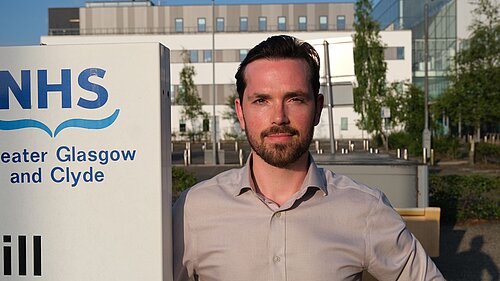 Adam Harley next to a sign with the NHS Greater Glasgow and Clyde logo with a hospital building in the background
