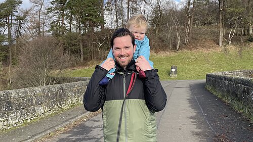 Adam Harley outside in a waterproof jacked with his son on his shoulders holding his hands.