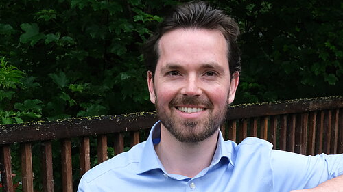 Adam Harley in a blue shirt standing with arm on top of a wooden fence of a bridge..