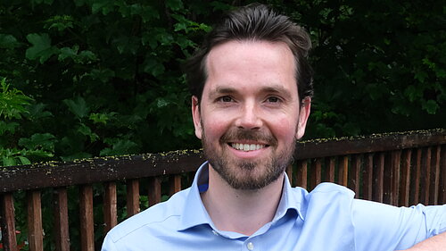 Adam Harley in a blue shirt standing with arm on top of a wooden fence of a bridge..